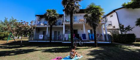 Un edificio residencial con balcón, césped y una persona sentada sobre una toalla.