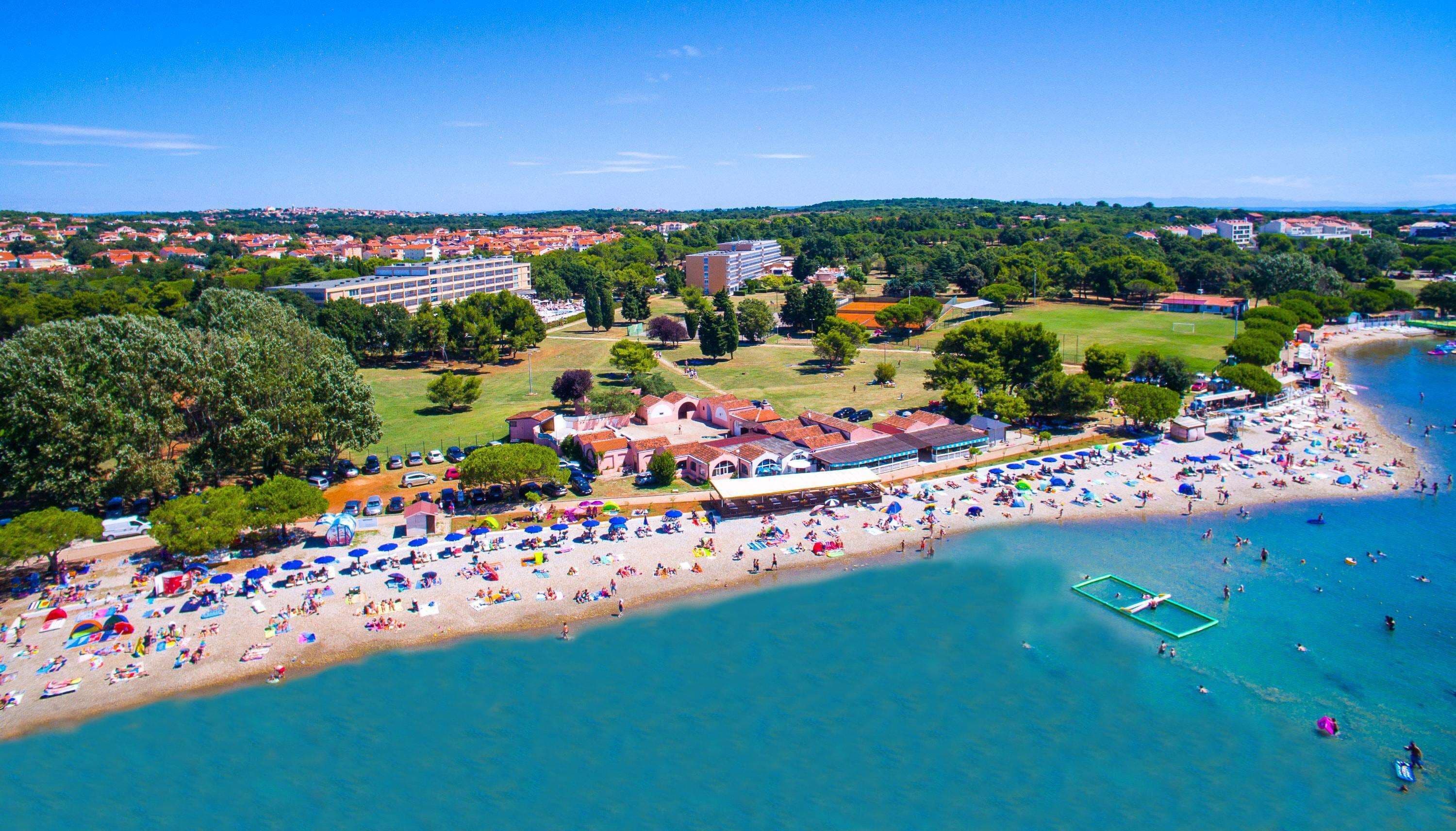 An aerial view of a coastal area with a beach, a swimming pool, and surrounding buildings.