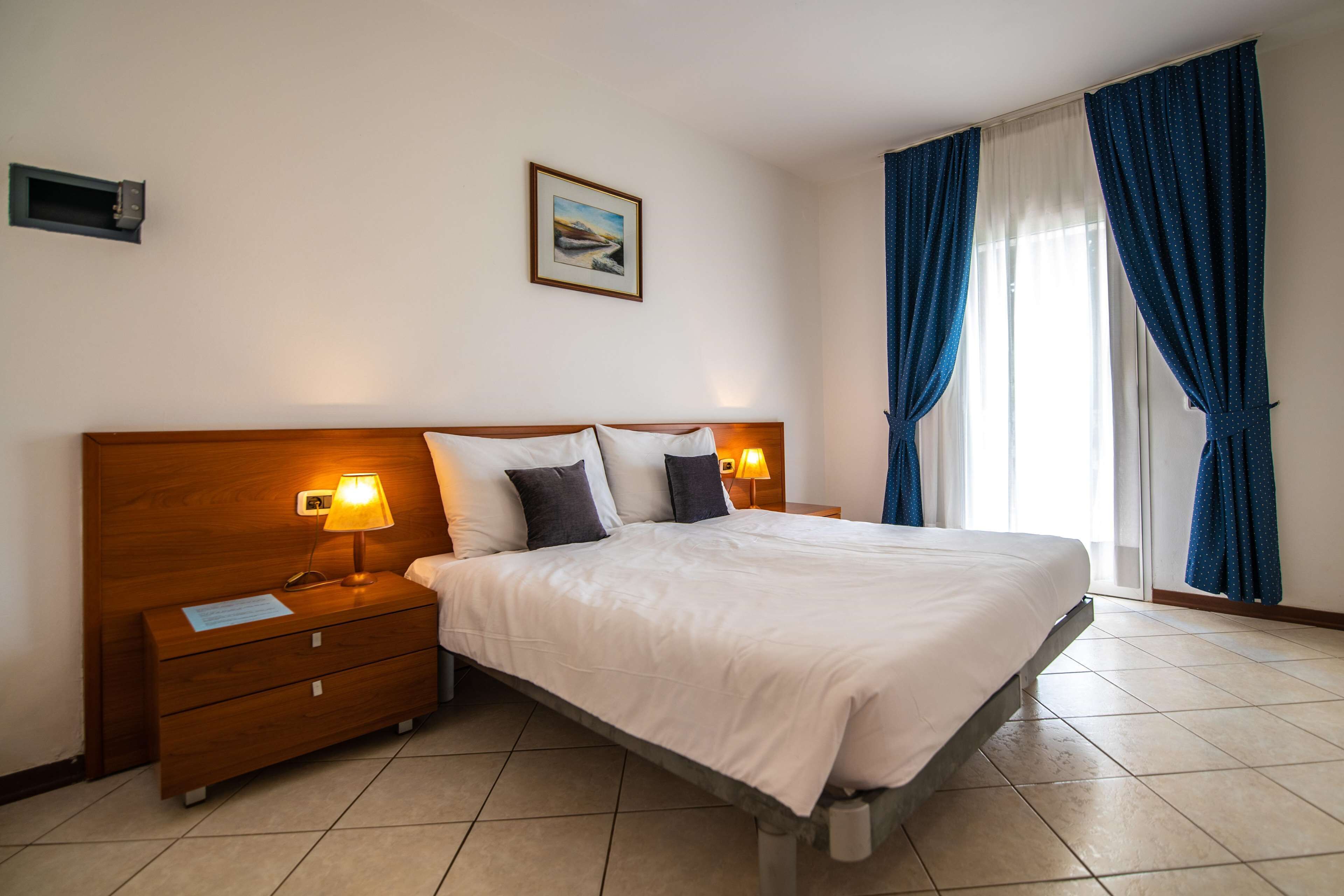 Habitación de hotel con una cama grande, dos mesitas de noche con lámparas, un atril y una ventana con cortinas. 