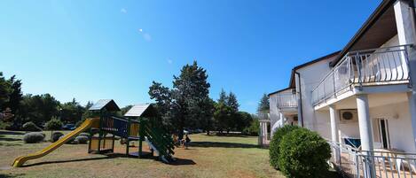 A playground with a slide and climbing structure, surrounded by grass and trees.