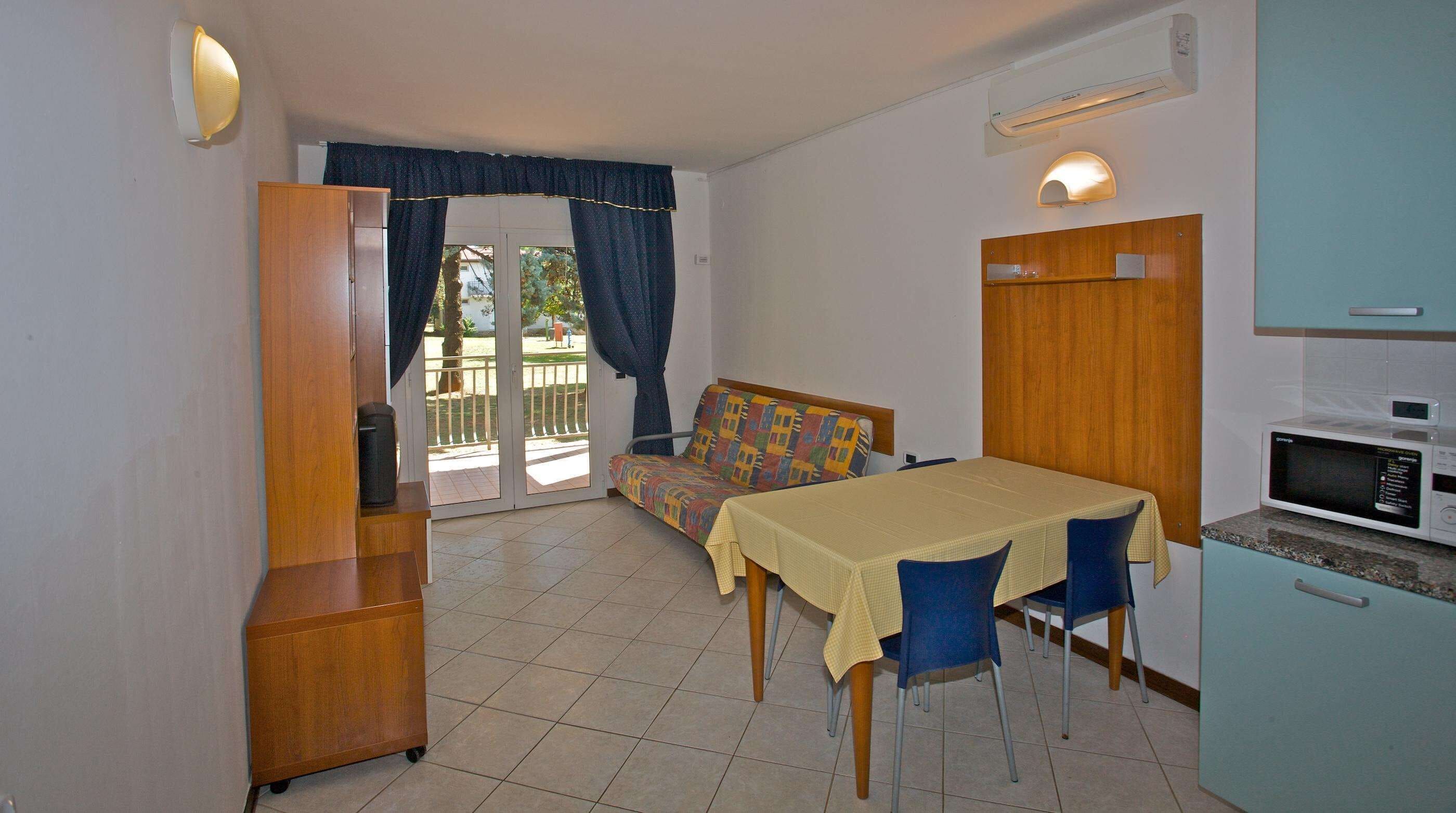 A compact room with a dining table, chairs, a sofa, a kitchenette, and a sliding glass door leading to an outdoor area.