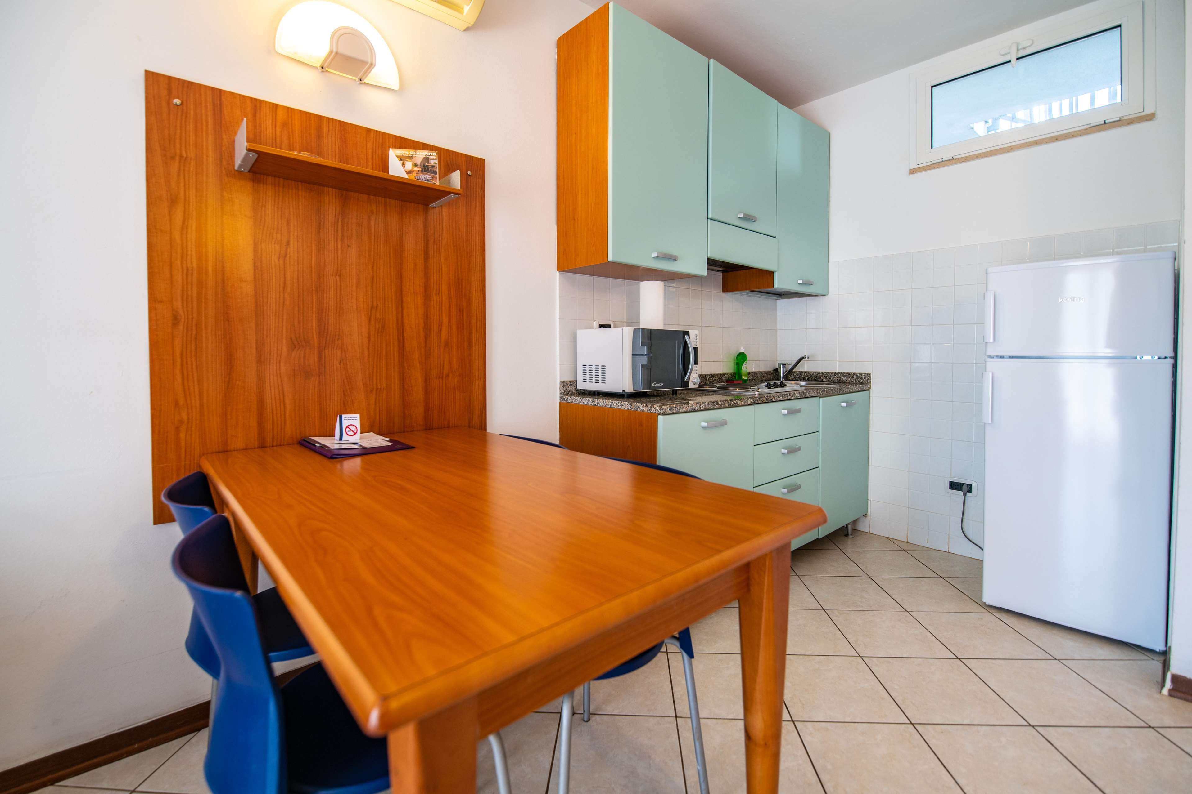 Una cocina con mesa de madera, sillas azules, microondas, refrigerador y armarios color turquesa.
