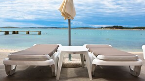 A beachside lounge area with two sunbeds and a table under a canopy, overlooking a calm sea and a distant shoreline. - Ai Pini Medulin Resort (Medulin)