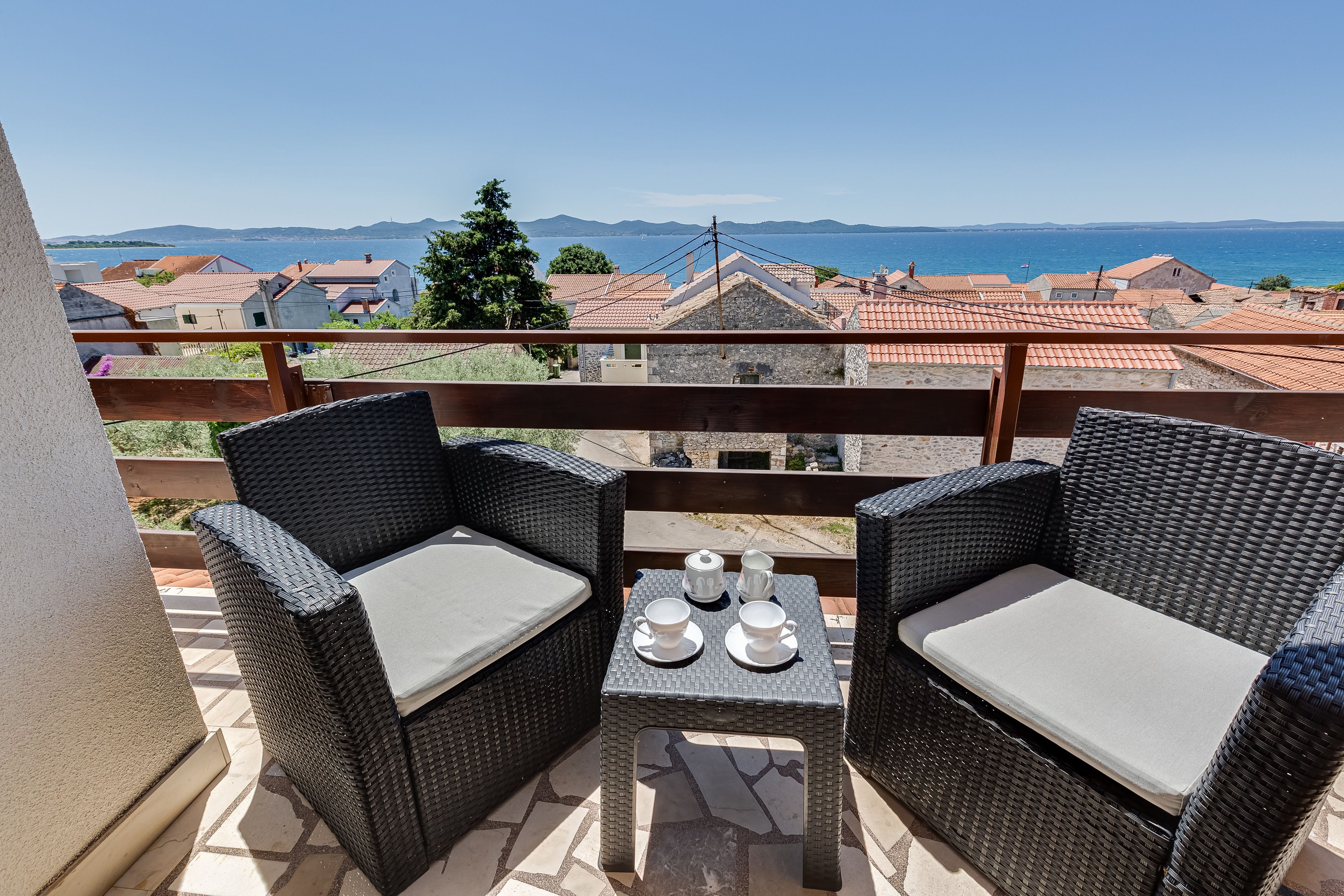 Een balkon met rieten meubels en een tafel gedekt voor twee, met uitzicht op een kustplaatsje.