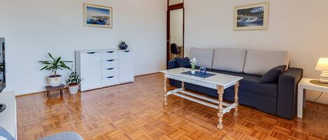 A living room with a sofa, a coffee table, a TV, and a kitchen area with cabinets and a plant.