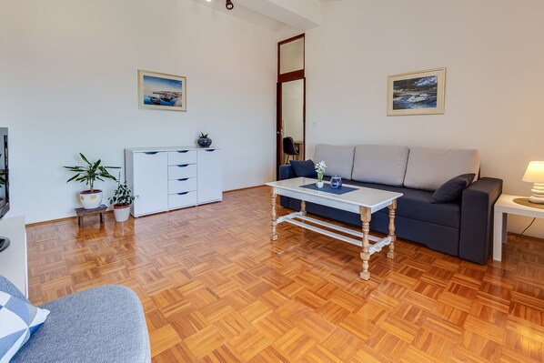 A living room with a sofa, a coffee table, a TV, and a kitchen area with cabinets and a plant. - Baresic Apartmani (Zadar)