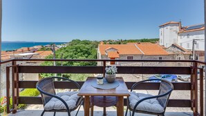 A balcony with wicker chairs, a table with a vase, and a view of the sea and buildings. - Baresic Apartmani (Zadar)