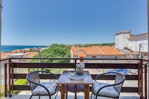 A balcony with wicker chairs, a table with a vase, and a view of the sea and buildings. - Baresic Apartmani (Zadar)