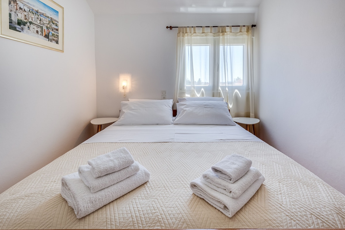 A hotel room with a bed, two bedside tables, a wall-mounted light, and a window with sheer curtains.