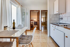 A modern kitchen with white cabinets, a dining area with a table and chairs, and a visible hallway leading to a bedroom. - Baresic Apartmani (Zadar)