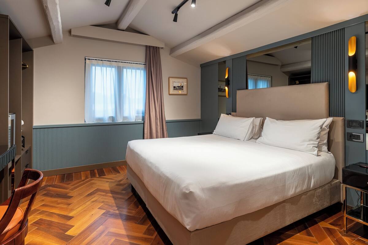 Une chambre avec un grand lit, un plancher en bois, une fenêtre avec des rideaux, un miroir et un luminaire fixé au mur.