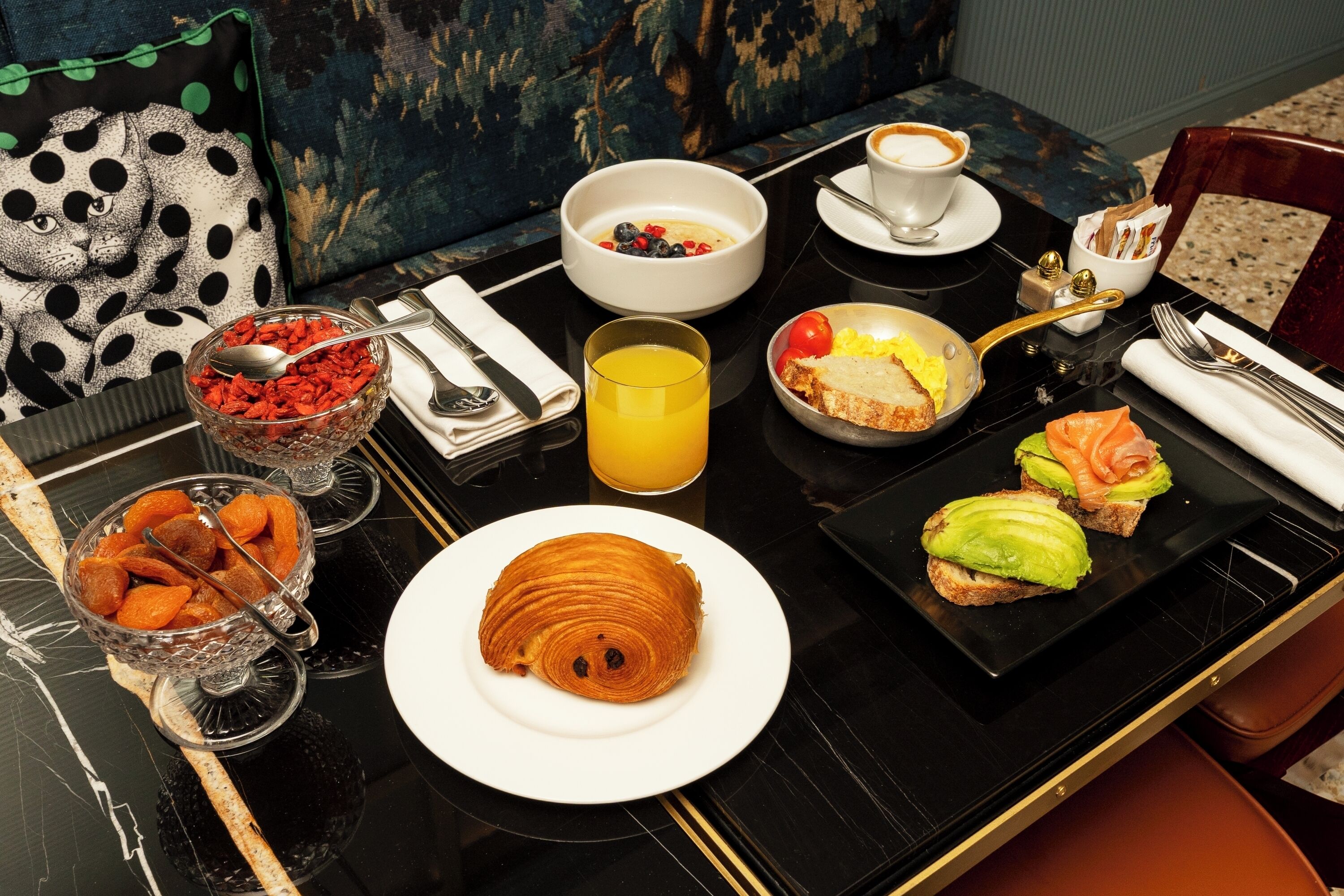 Una escena de desayuno con un croissant, tazones de bayas y frutas secas, un vaso de jugo de naranja y sándwiches sobre una mesa negra.