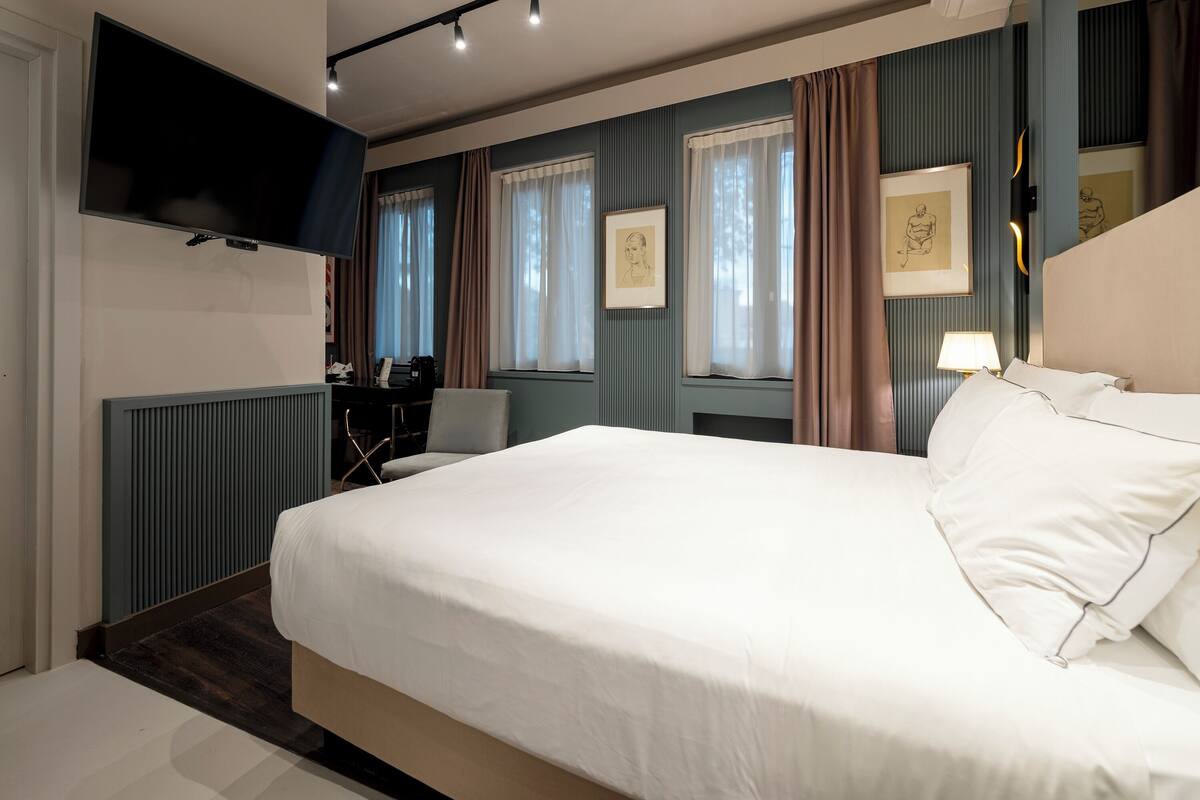 Une chambre d’hôtel dotée d’un grand lit, d’un téléviseur à écran plat fixé au mur, d’un bureau avec une chaise et d’une fenêtre avec des rideaux.