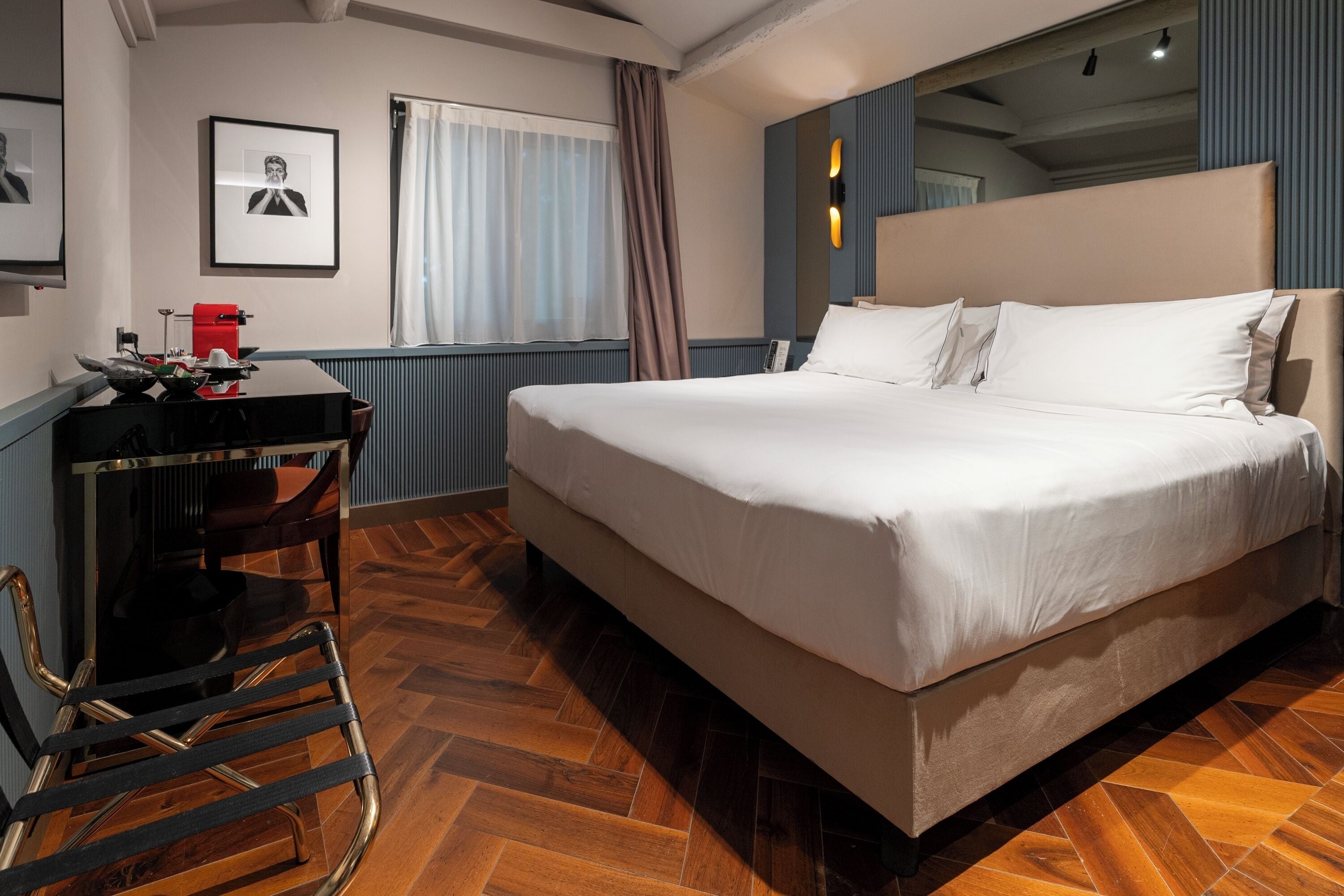 A hotel room with a large bed, a desk with a chair, a window with curtains, and a framed picture on the wall.