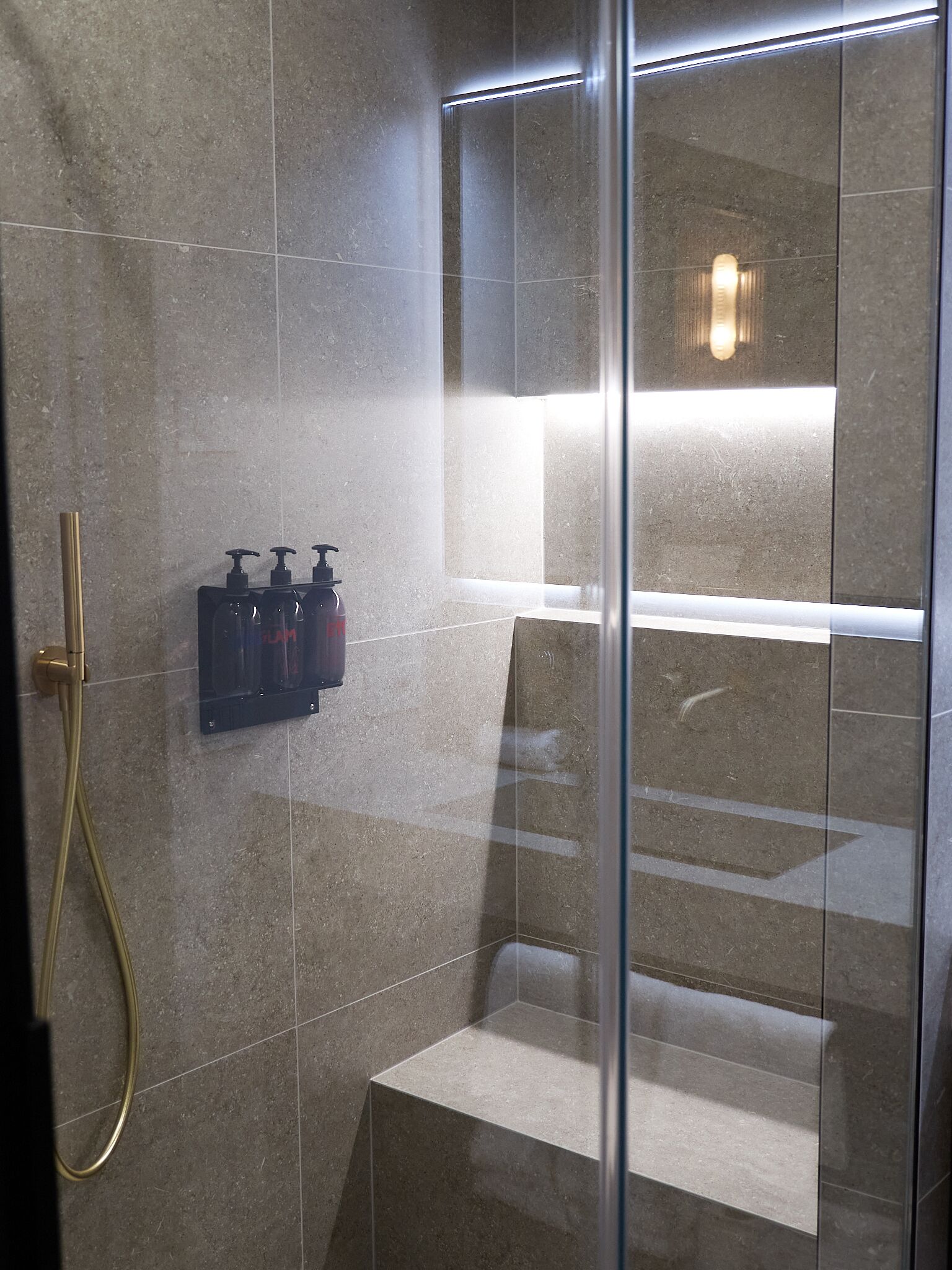 A modern shower area with a glass partition, a handheld showerhead, and two soap dispensers mounted on the wall.