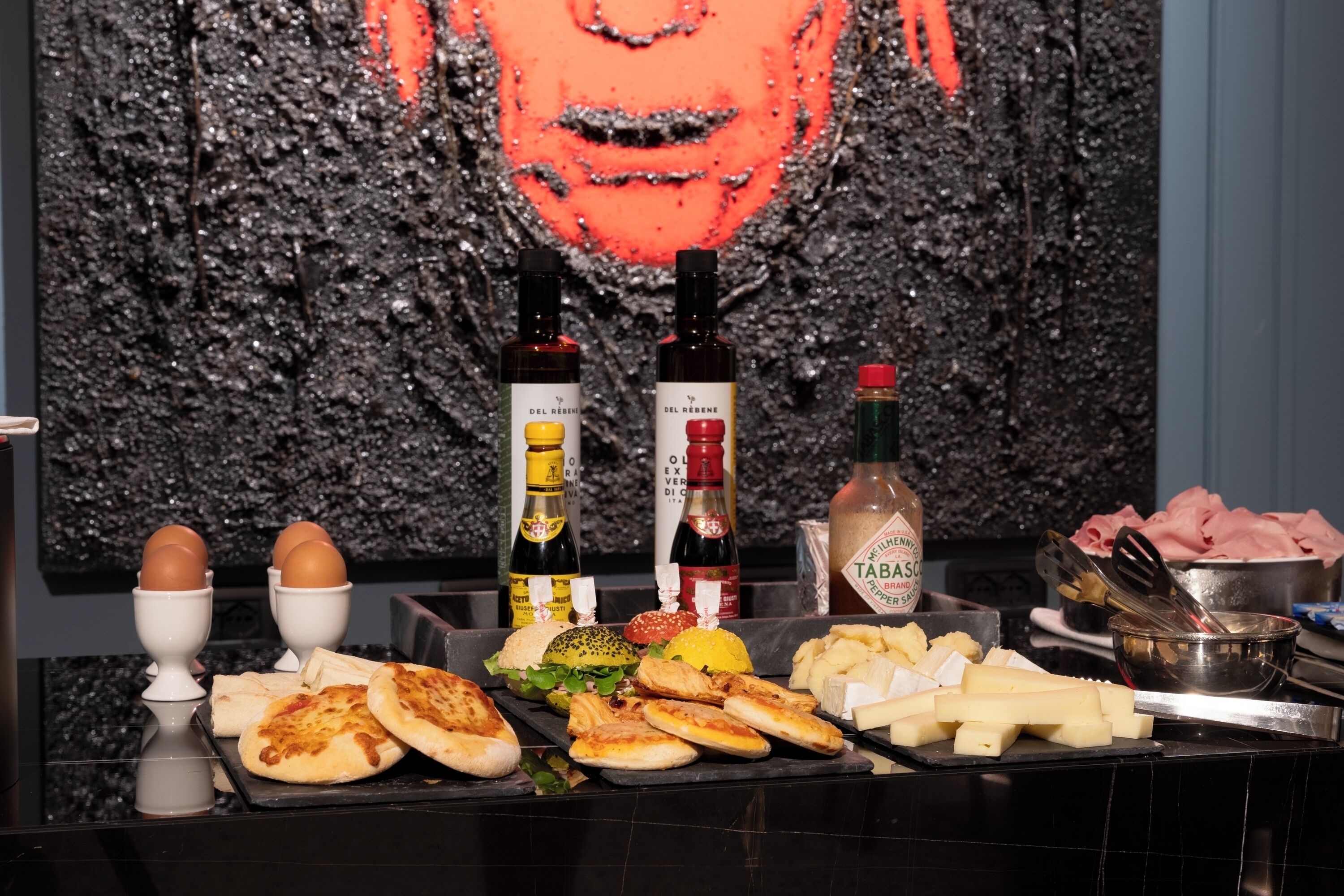 A buffet table with various food items, including eggs, bread, and meats, set against a textured wall with a large portrait.