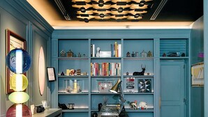 Een kamer met een blauwe wand, een plafond met een uniek patroon, een bureau met een lamp en een boekenplank met diverse spullen.