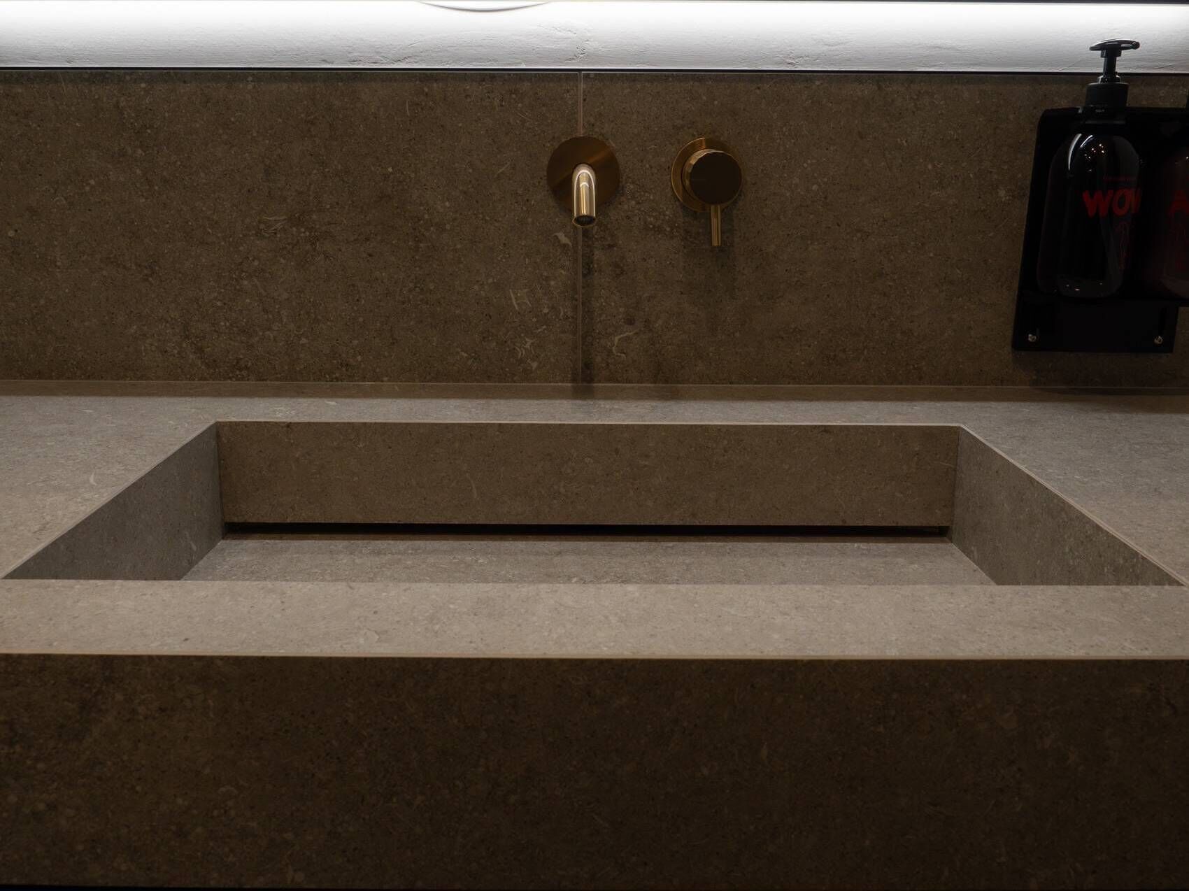 A bathroom with a rectangular sink, two gold-colored faucets, and a soap dispenser on the wall.