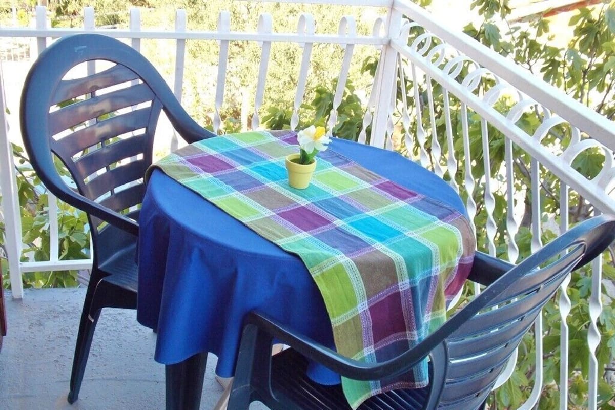 A round table with a plaid tablecloth and a potted plant on a balcony with white railings and green foliage in the background.