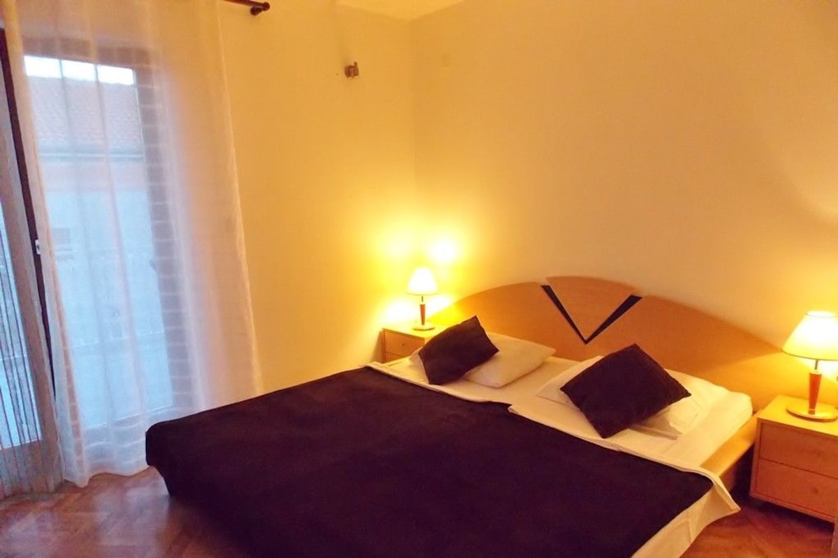 A hotel room with a bed, two bedside lamps, a wooden headboard, and a window with sheer curtains.