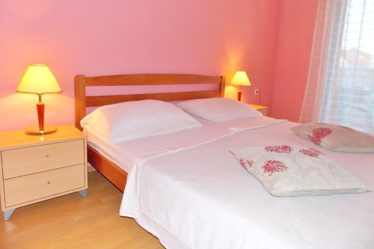 A bedroom with a wooden bed, two bedside tables with lamps, and a window with sheer curtains.