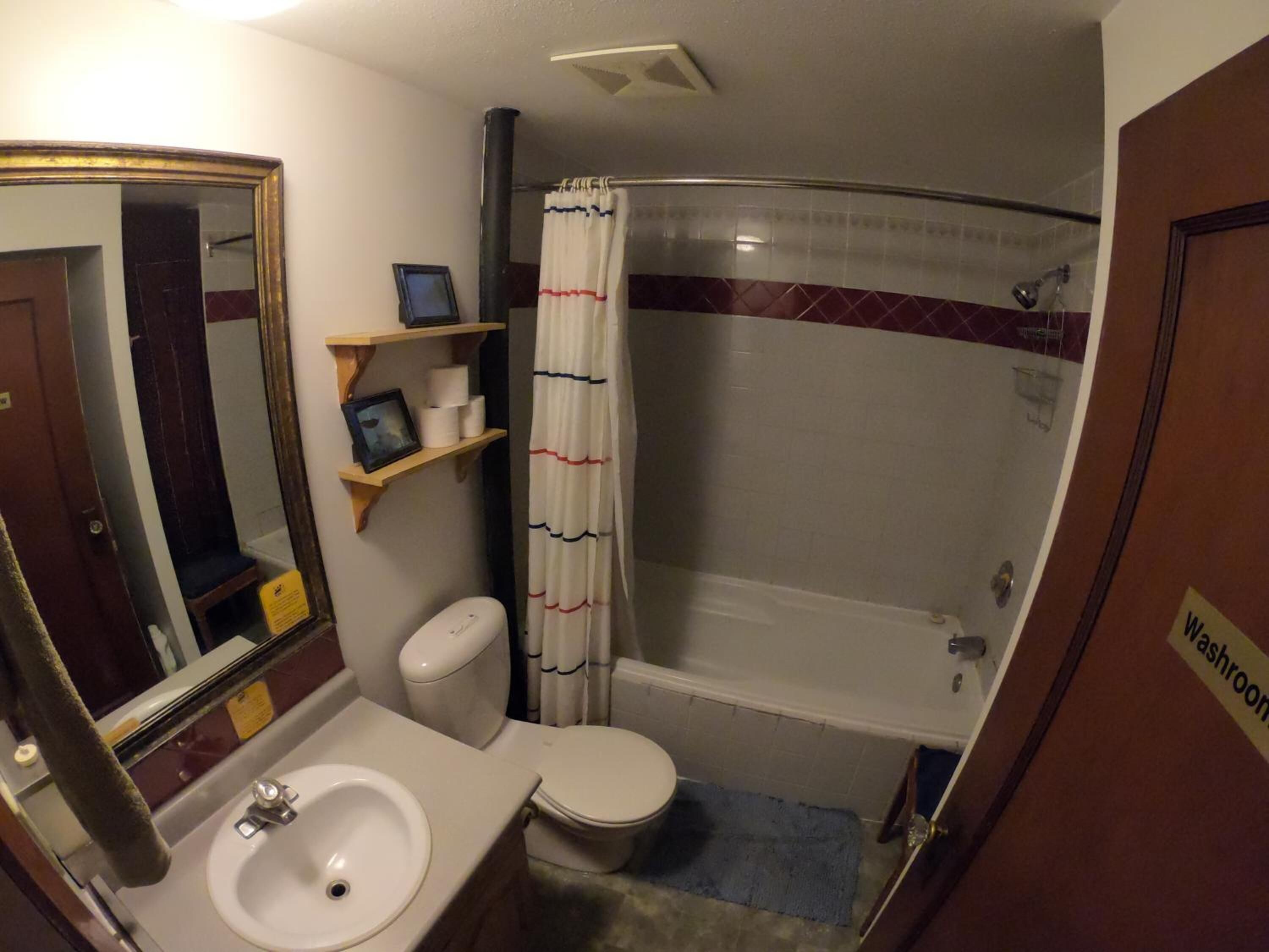 Un baño compacto con inodoro, lavamanos y ducha. Se ve un espejo y una repisa con artículos de aseo.