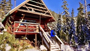 Une cabane en bois avec un escalier raide menant à une plateforme, le tout entouré d’arbres couverts de neige.