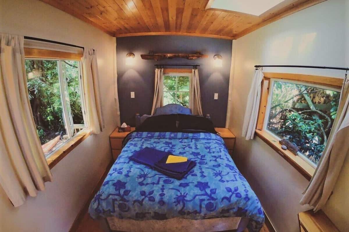 Een slaapkamer met een bed, twee ramen en uitzicht op bomen.