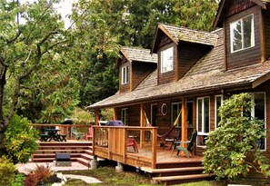 A wooden cabin with a porch, outdoor seating, and a small deck. - Up The Creek Backpacker's Lodge (Roberts Creek)