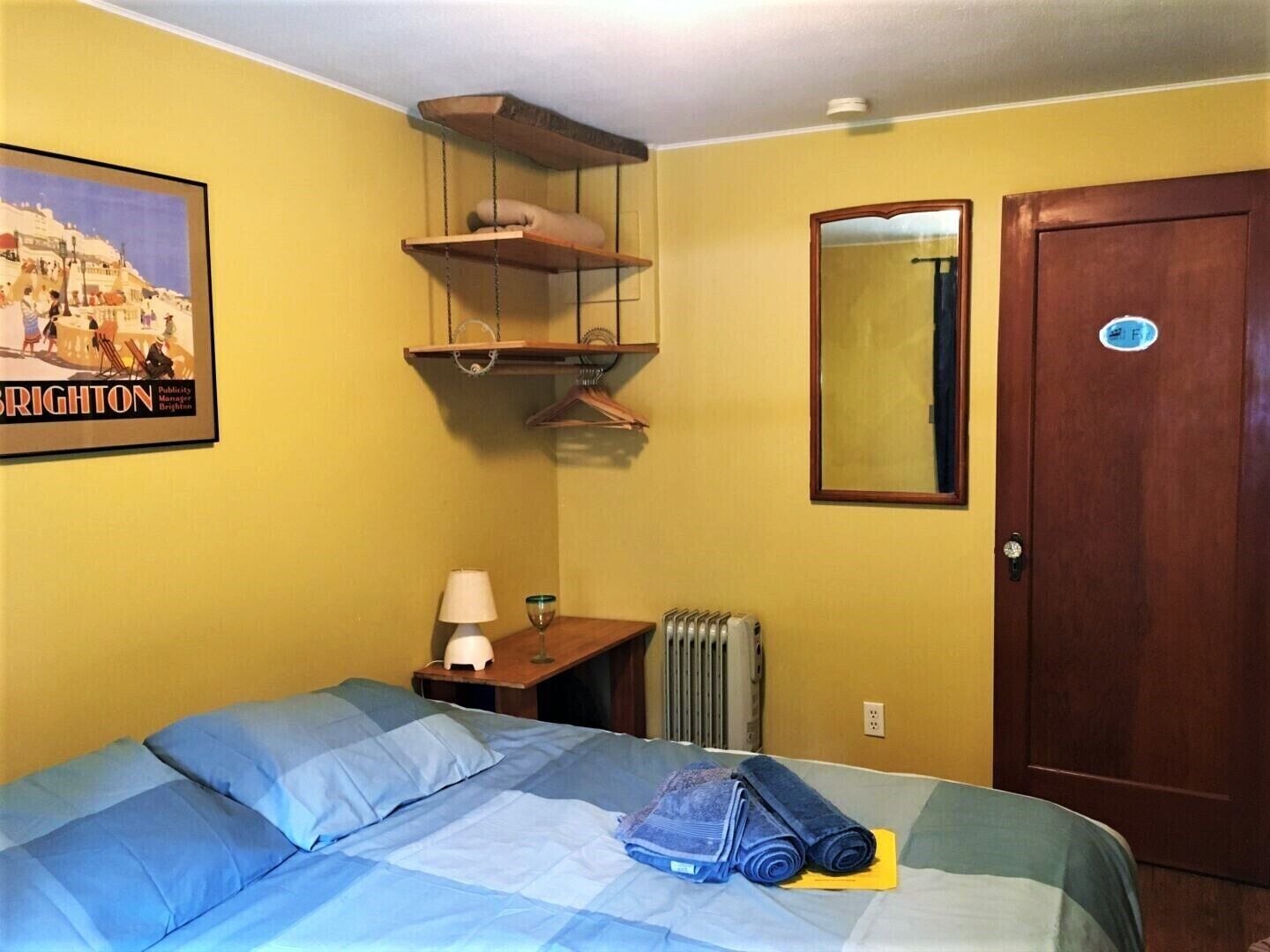 Une pièce comprenant un lit, une petite table avec une lampe, un radiateur, une étagère en bois et une porte.