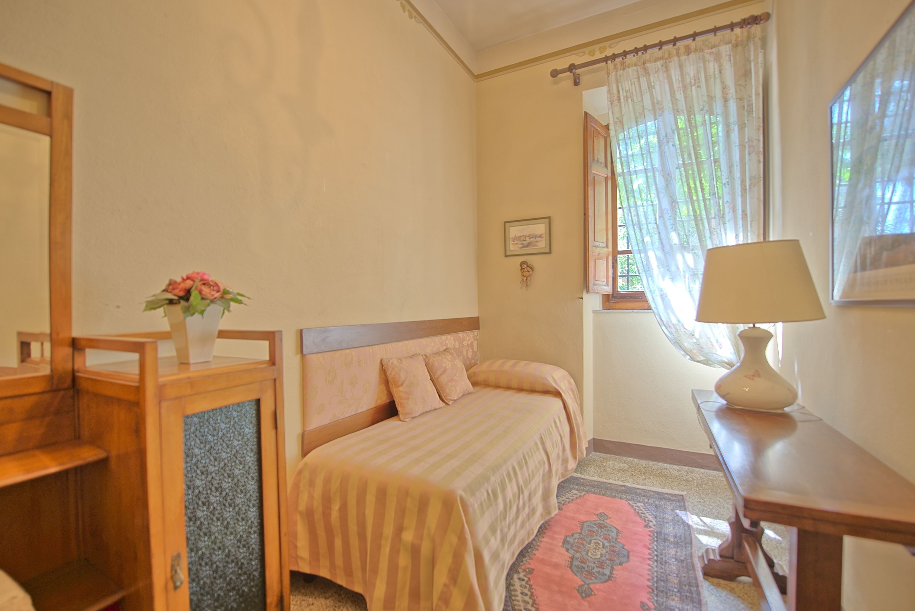 Une chambre à coucher avec un lit, une table de chevet avec une lampe, une armoire en bois, une fenêtre avec des rideaux et un tableau encadré au mur.