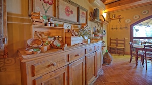 Une cuisine rustique avec des armoires en bois, un plan de travail en bois et divers ustensiles de cuisine. Les murs sont ornés de tableaux représentant des motifs floraux encadrés, et une fenêtre offre une vue sur l’extérieur.