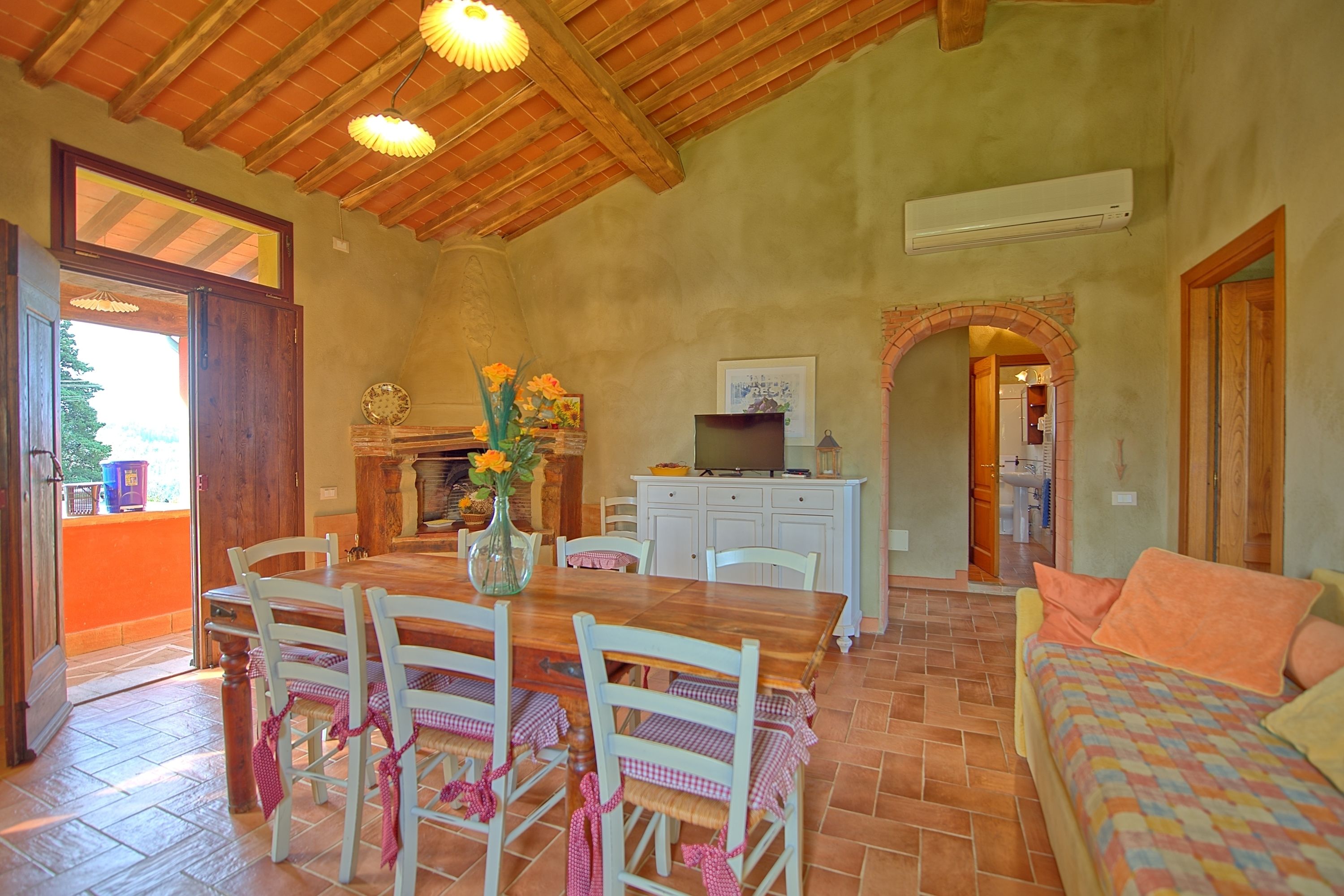 A dining area with a wooden table and chairs, a vase with flowers, and a kitchen area with cabinets and an air conditioning unit.