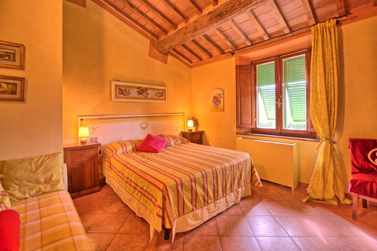 Un dormitorio con cama, mesitas de noche, una ventana con contraventanas y una silla roja.