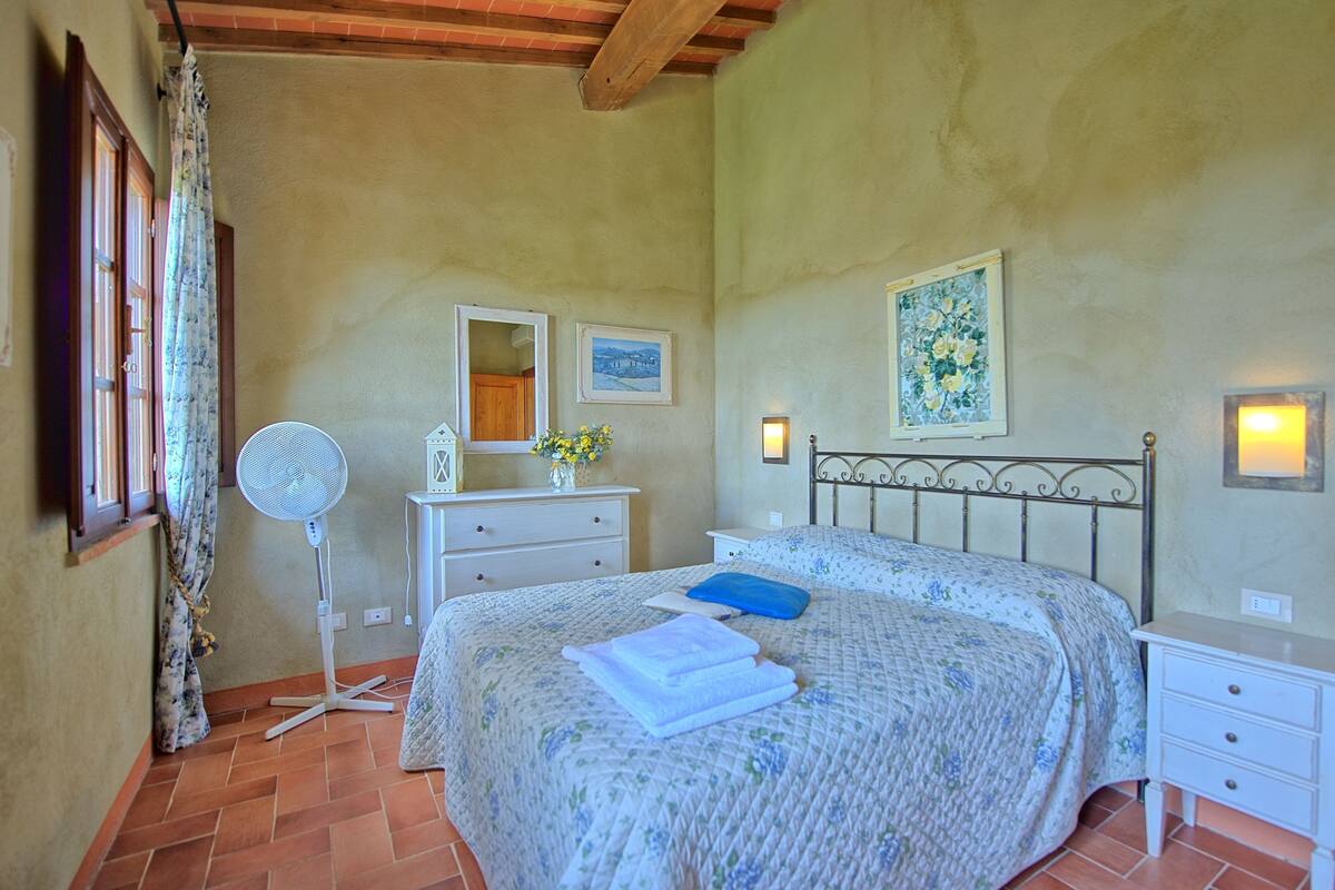 Un dormitorio con cama, mesita de noche, cómoda, ventilador y ventana con cortinas.