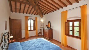 A bedroom with a wooden ceiling, a bed with a blue quilt, a dresser, and two windows with yellow curtains. - Certaldo (Certaldo)