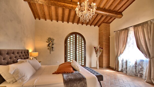 A bedroom with a large bed, wooden ceiling beams, a chandelier, and a view of the outdoors through a window with curtains. - Villa Nora (Empoli)
