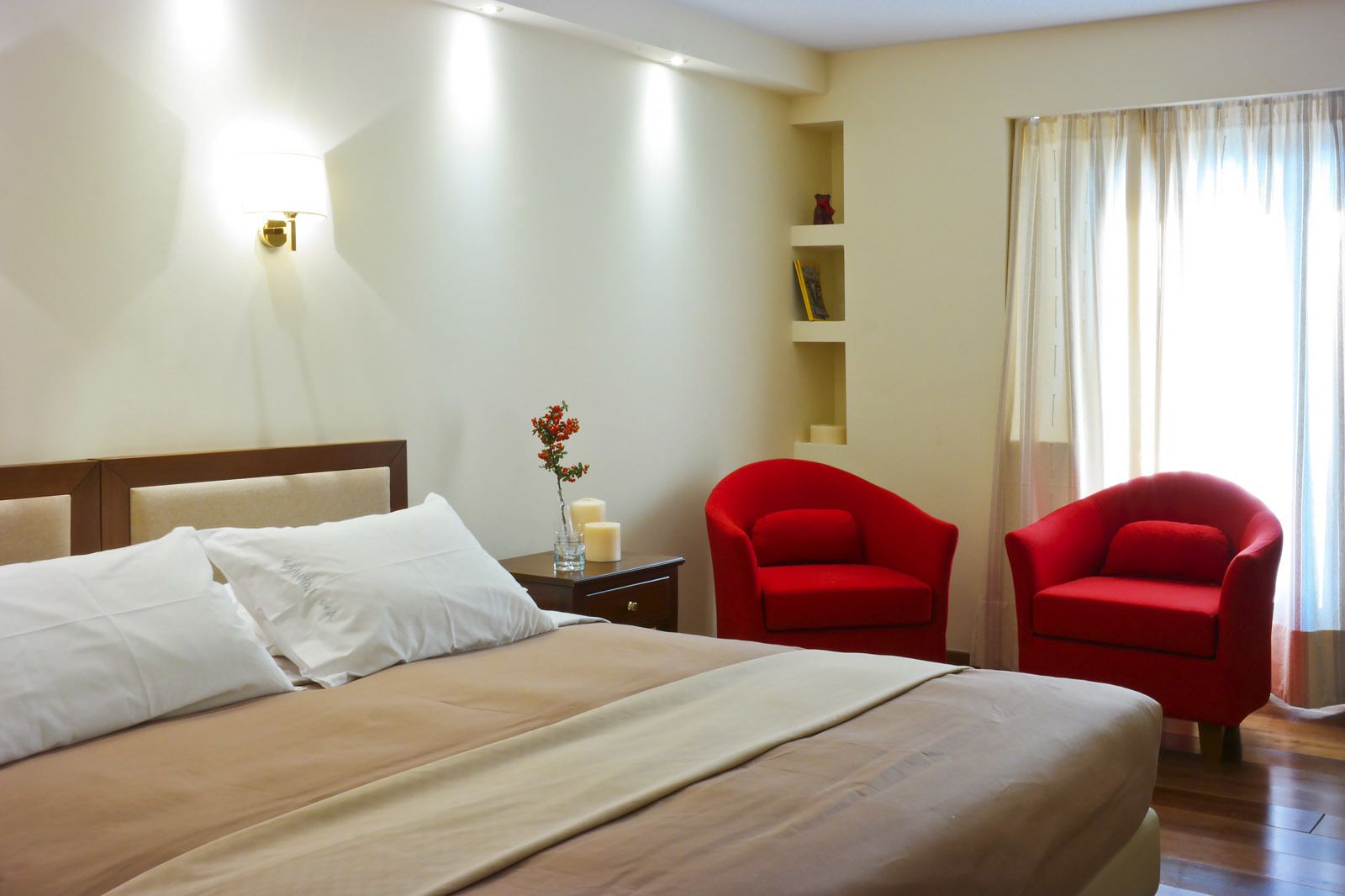 Una camera d'albergo con un letto grande, due poltrone rosse, un comodino con un vaso di fiori e una lampada a parete.
