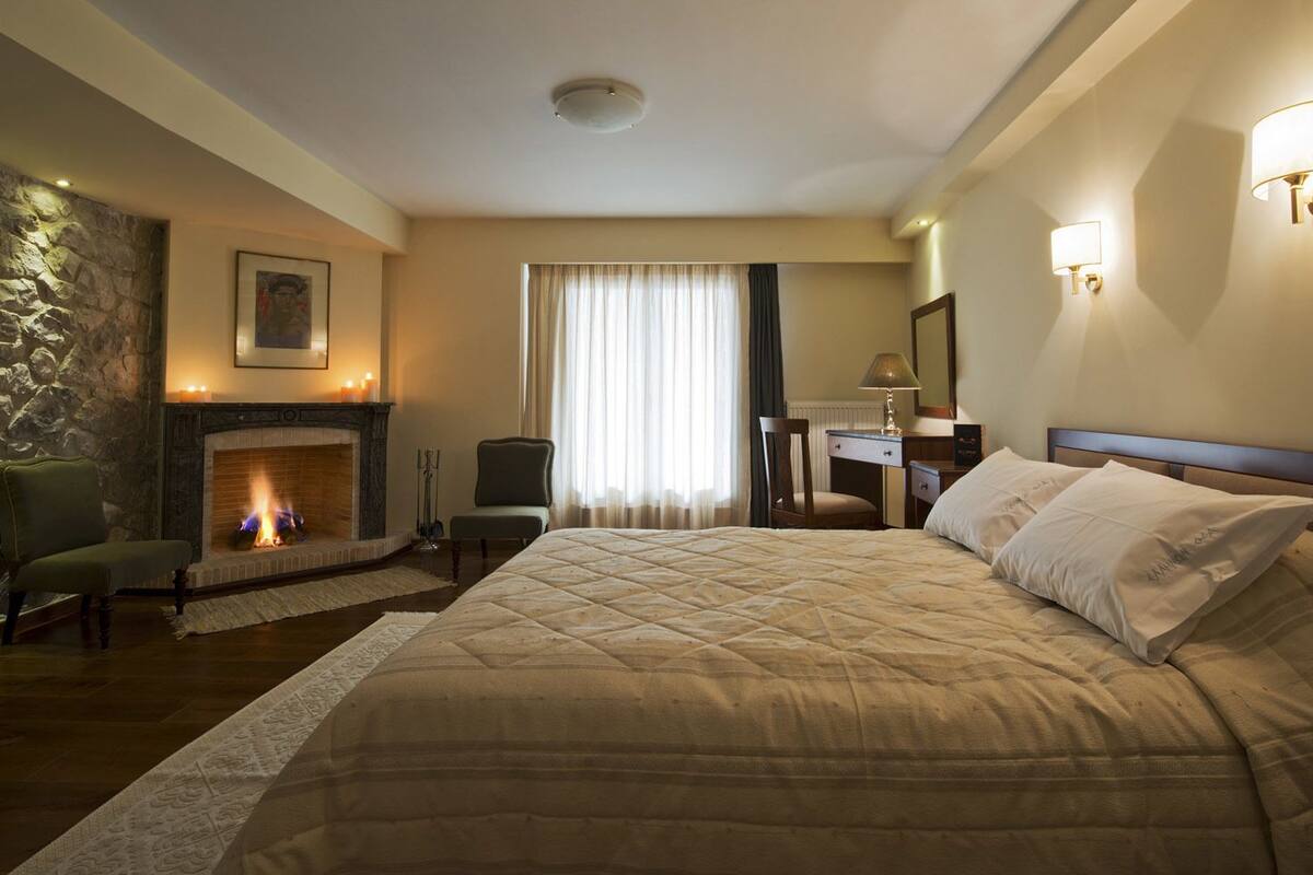 Une chambre d’hôtel avec un grand lit, une cheminée, un bureau avec une chaise et une fenêtre avec des rideaux.