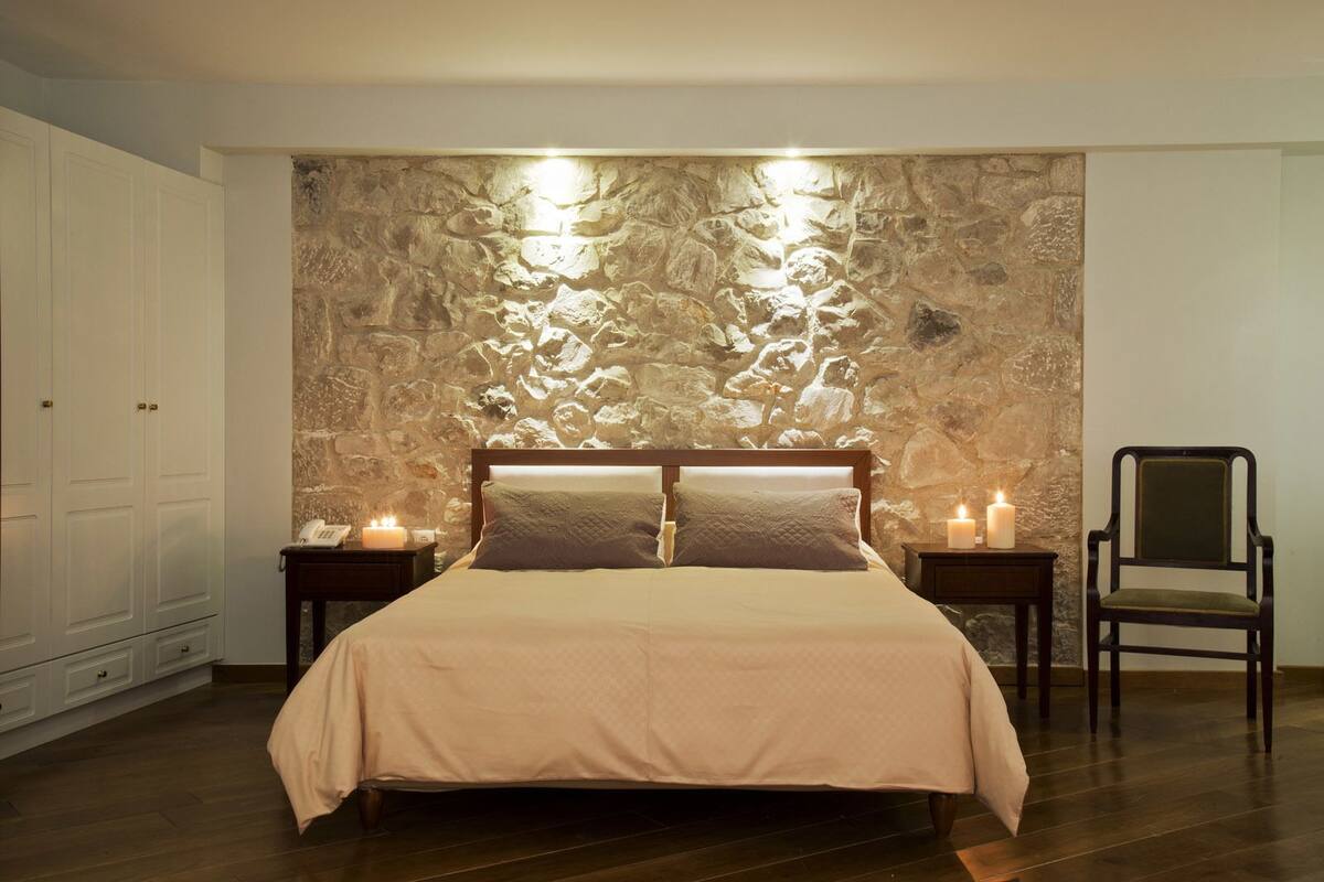 Une chambre avec un mur en pierre, un lit avec une tête de lit en bois, deux tables de chevet avec des bougies, une chaise et une armoire.