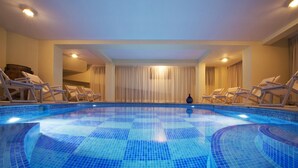Uma grande piscina interior com chão e paredes azulejadas em azul, rodeada por espreguiçadeiras e cortinas.