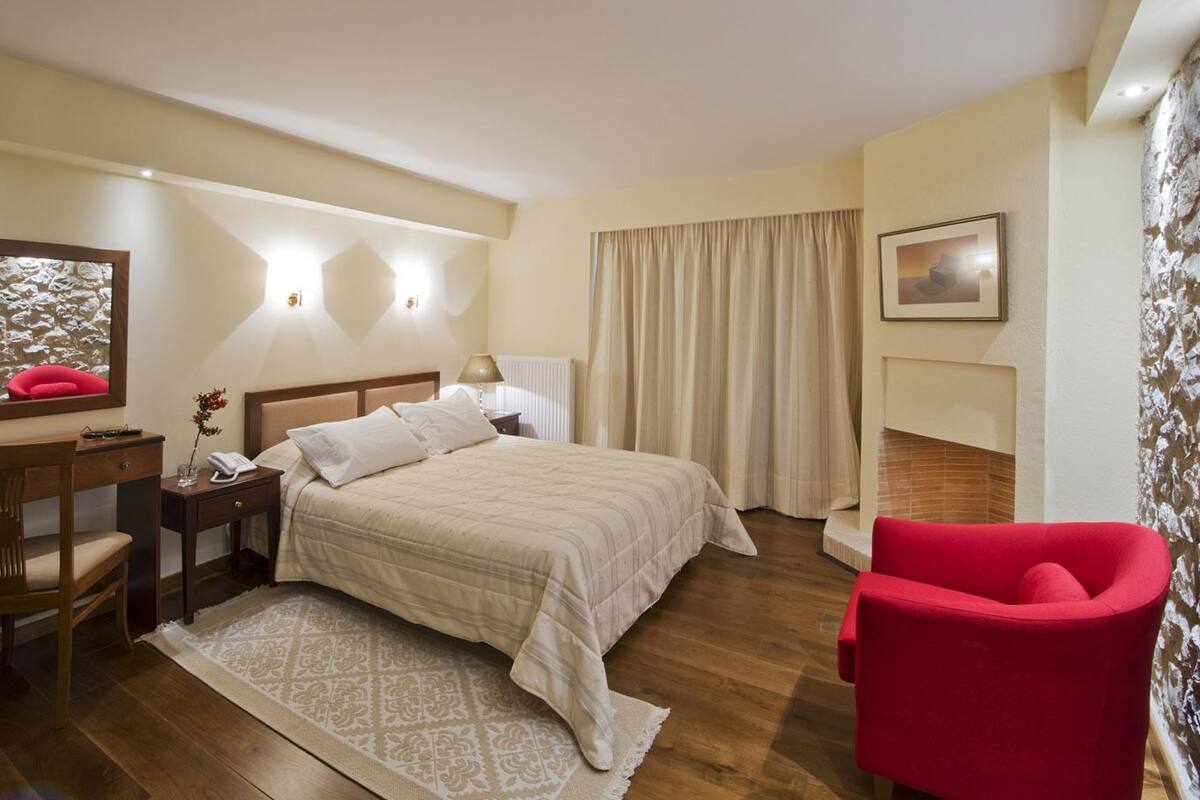 Une chambre d’hôtel avec un lit, un bureau, une chaise, un fauteuil rouge, un tableau et une cheminée.