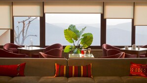 Quarto com sofá, duas poltronas, uma mesa com planta e vista para montanhas através de amplas janelas.