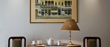A framed historical street scene painting above a small round table with a tablecloth, two chairs, and a table lamp.