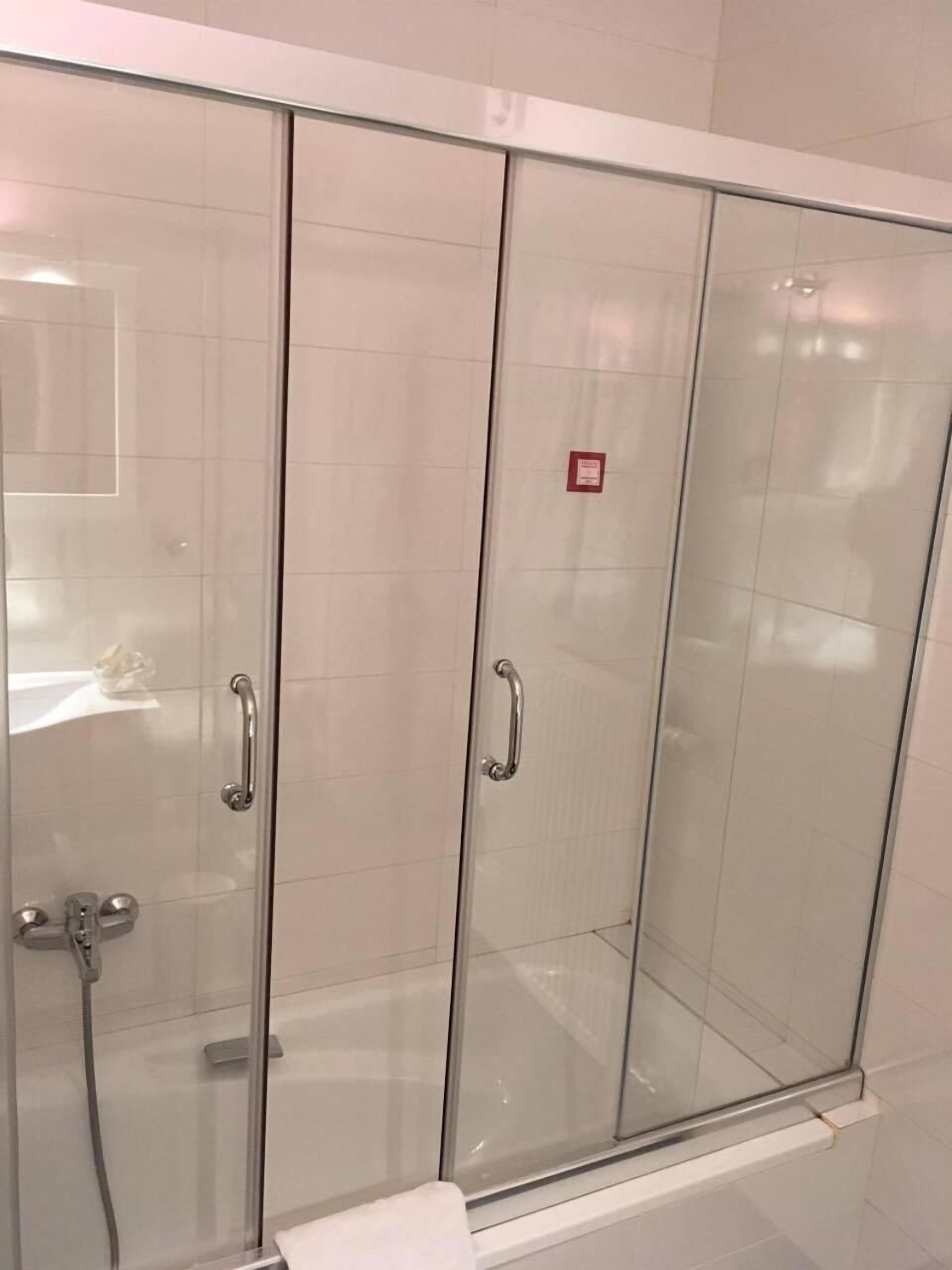 A glass-enclosed shower stall with a white tiled wall, a handrail, and a small red sign on the door.