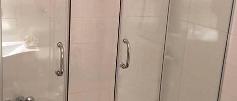 A glass-enclosed shower stall with a white tiled wall, a handrail, and a small red sign on the door.