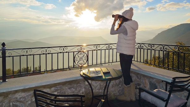 Uma pessoa fotografando uma vista panorâmica de montanhas a partir de uma varanda com mesa e cadeiras.