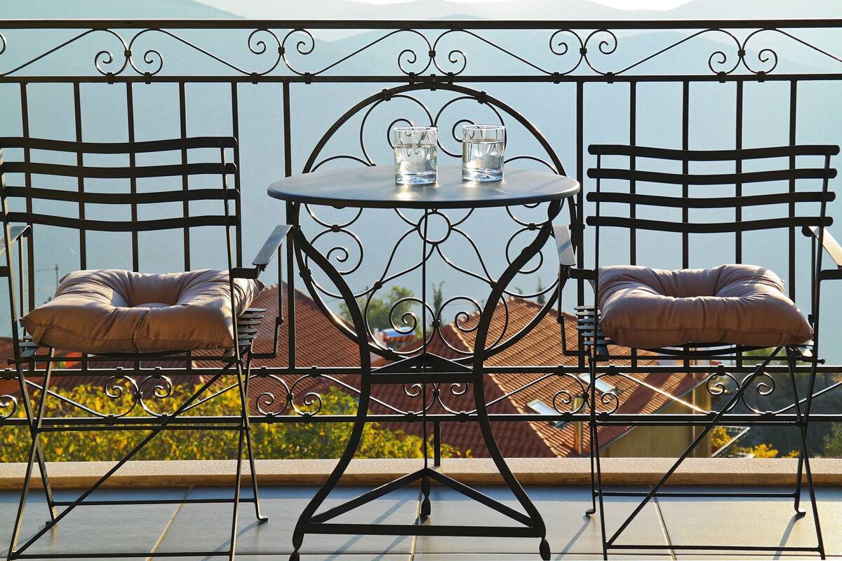 Un balcon agrémenté d’une table en fer forgé et de deux fauteuils rembourrés, d’où l’on peut admirer un immeuble avec un toit rouge.