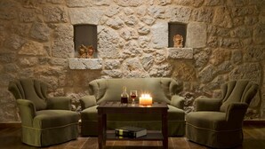 Um quarto acolhedor com parede de pedra, dois poltronas verdes, uma mesa de centro de madeira com uma vela, uma garrafa e um copo.