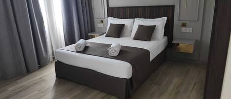 A modern hotel room with a large bed, dark curtains, and a wooden floor.